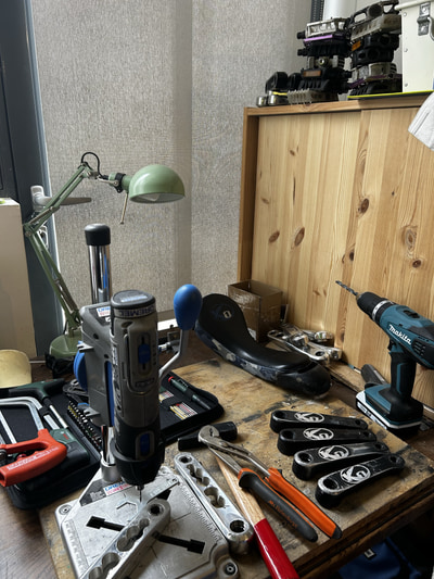 Établi de réparation de monocycles en bois. Sur le plan de travail sont disposés de nombreux outils. Une selle de monocycle noire repose au centre de l’établi, à côté de plusieurs manivelles noires.
